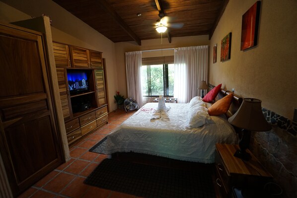 A bedroom with a bed, a TV, wooden cabinets, a ceiling fan, and a window with curtains.