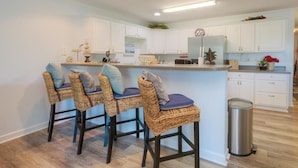 A kitchen with white cabinets, a stainless steel refrigerator, and a bar area with wicker chairs. - Stunning oceanview condo in Oak Island, perfect for families up to 8! (Oak Island)