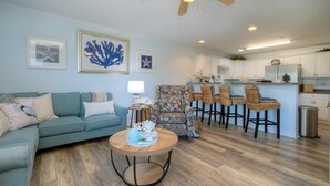 A living room with a sofa, a round coffee table, a patterned armchair, and a dining area with a table and chairs. - Stunning oceanview condo in Oak Island, perfect for families up to 8! (Oak Island)