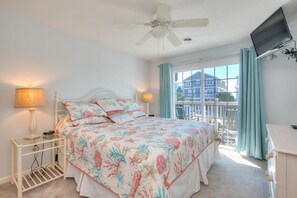 A bedroom with a bed, a ceiling fan, a TV, a window with curtains, and a balcony.