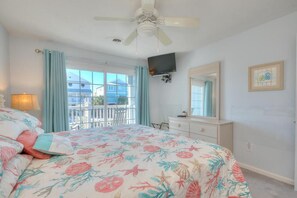 A bedroom with a bed, a ceiling fan, a TV, a dresser, a mirror, and a balcony view.