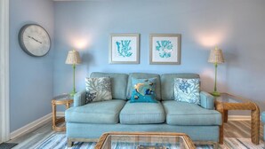 Uma sala de estar com um sofá azul, duas mesas laterais com luminárias, um relógio e obras de arte emolduradas na parede.