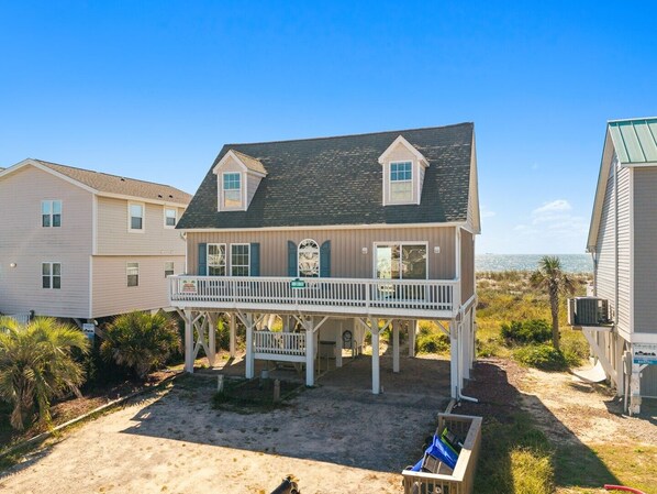 A two-story beach house with a balcony, a sandy driveway, and a view of the ocean. - Direct Oceanfront Home | Sleeps 12 with Stunning Views | Atlantic Breeze (Oak Island)