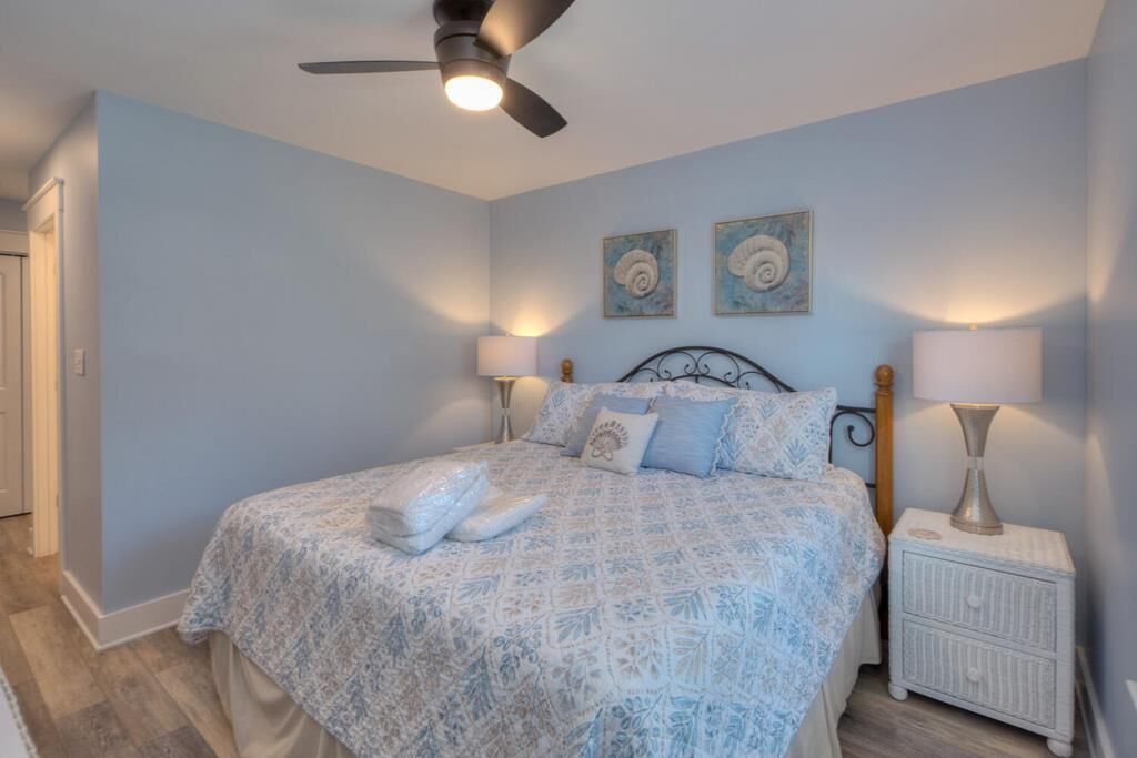 A bedroom with a bed, two wall-mounted lamps, a ceiling fan, and two framed artworks on the wall.