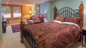 Un dormitorio con una cama grande, cabecera de madera, colcha con estampado rojo, lámpara roja y vista a un paisaje nevado por la ventana.