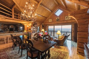 A spacious log cabin interior with a dining area, a chandelier, a fireplace, and a large window offering a view.