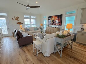 A spacious living room with a large television, a ceiling fan, and a view of the outdoors through the windows.