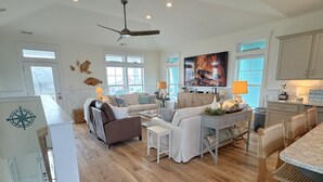A spacious living area with a large TV, wooden flooring, and a ceiling fan. - A Toast at the Coast: Luxury oceanview home in Oak Island, featuring 5 kings, private heated pool. (Oak Island)