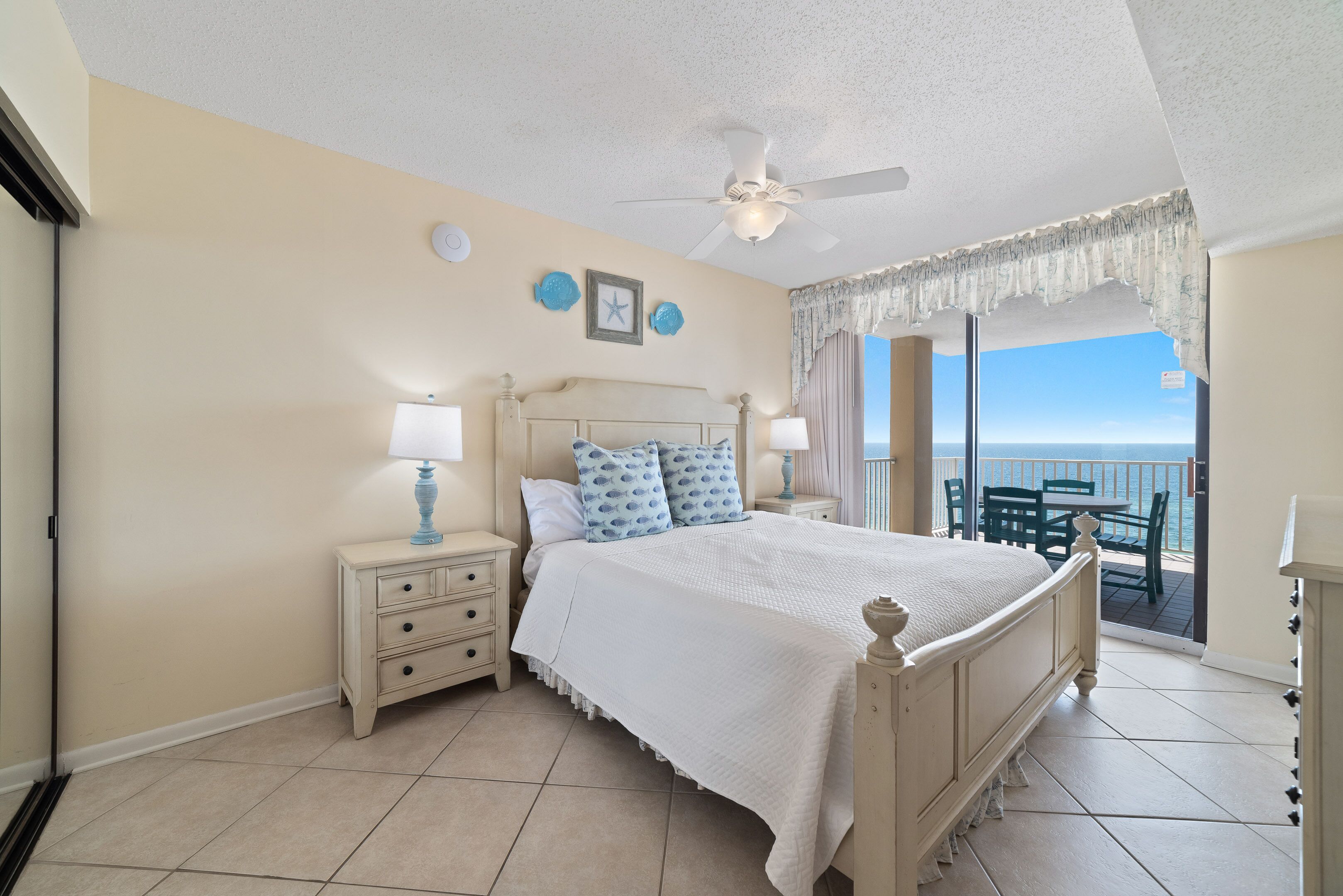 A bedroom with a bed, nightstands, a ceiling fan, and a view of the ocean through a sliding door.
