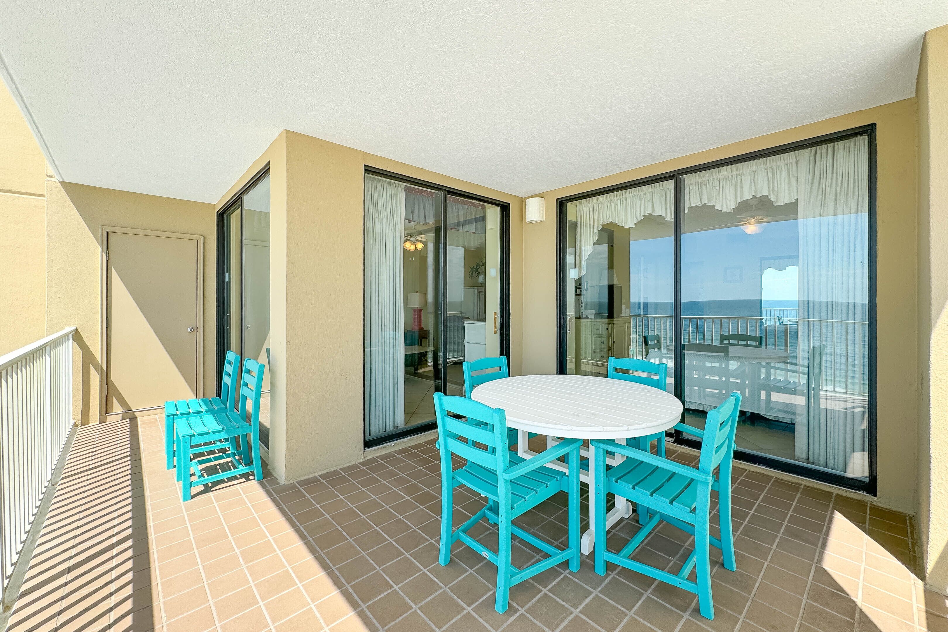 Un balcon avec une table blanche et des chaises turquoise, de grandes portes vitrées et une vue sur la mer.