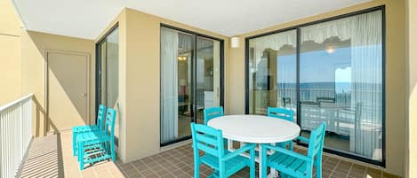 Un balcon avec une table blanche et des chaises turquoise, de grandes portes vitrées et une vue sur la mer.