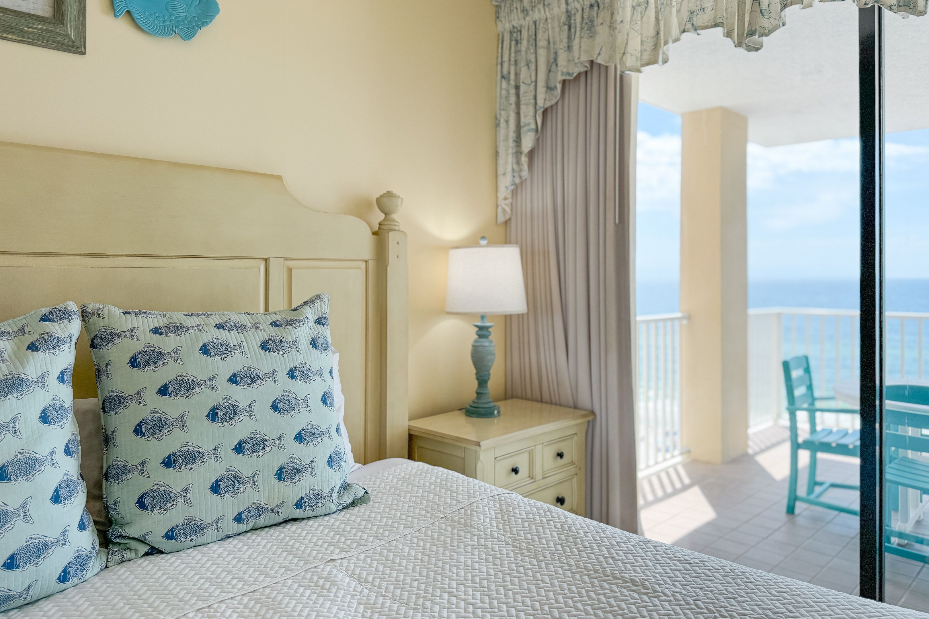 A bedroom with a bed, pillows, a nightstand, a lamp, and a view of the balcony with chairs.