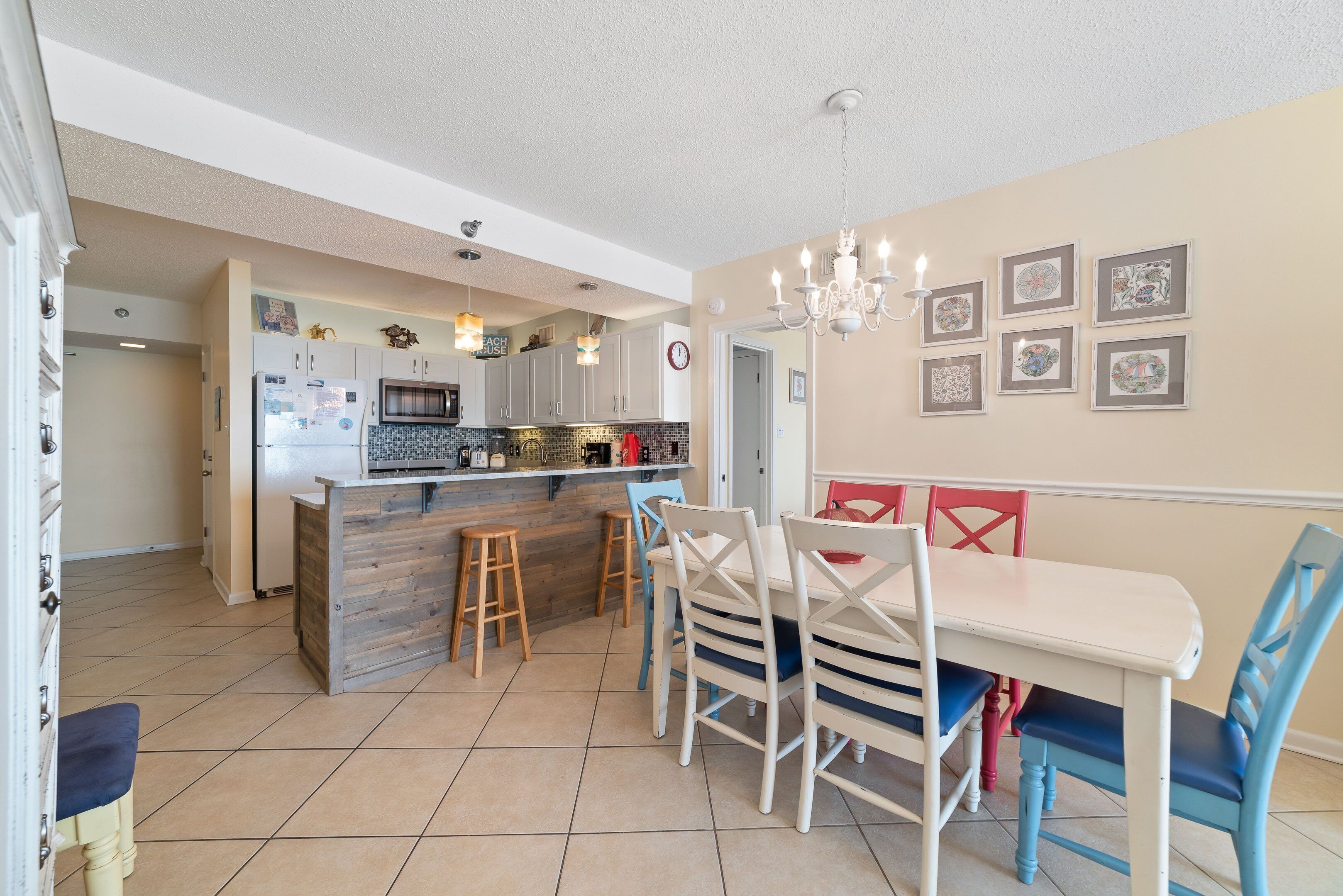 A modern kitchen with a central island, wooden stools, a dining table with chairs, and a chandelier.