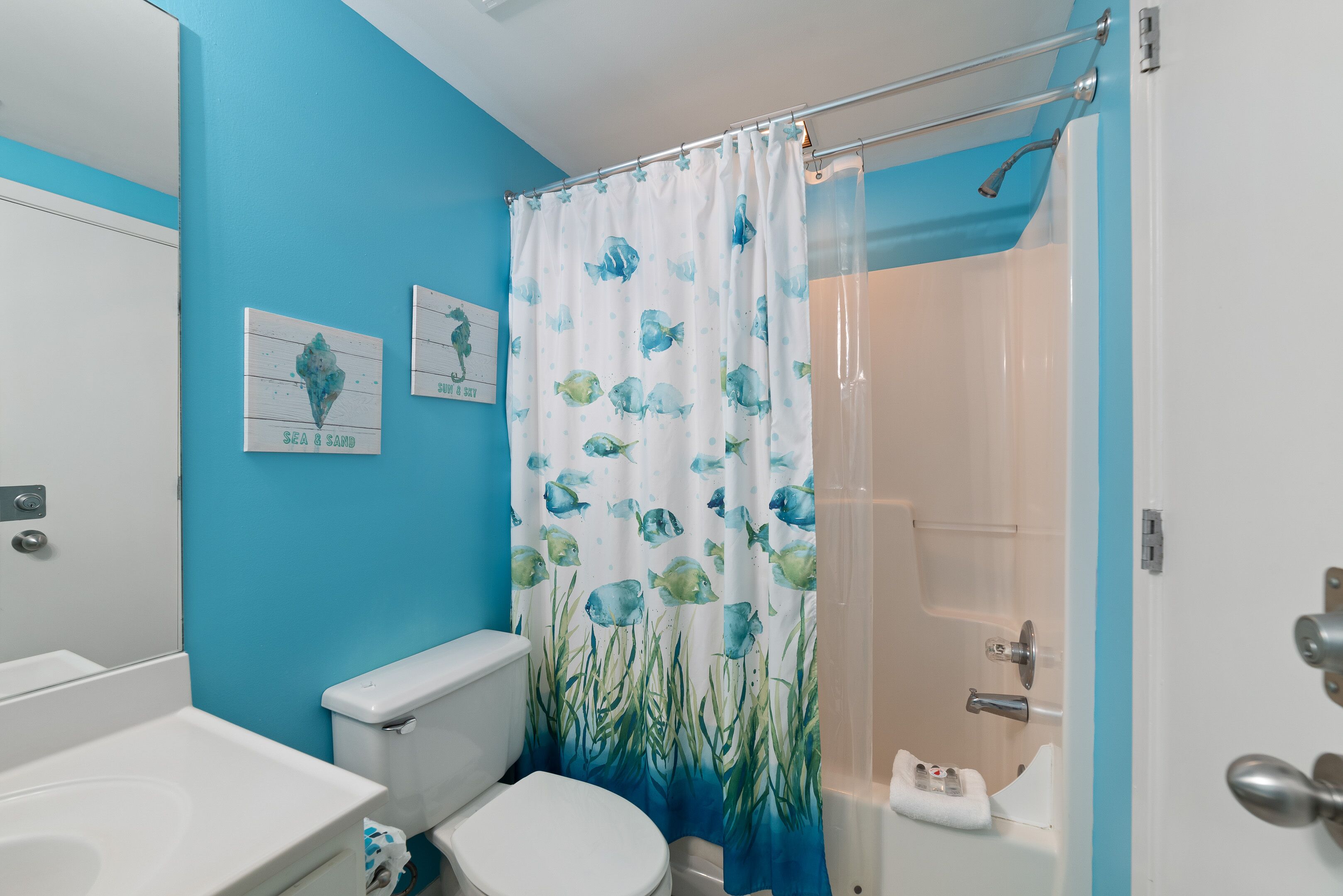 Een badkamer met een blauwe wand, een wit toilet, een witte wastafel en een douchegordijn met een ontwerp van vissen en zeewier.