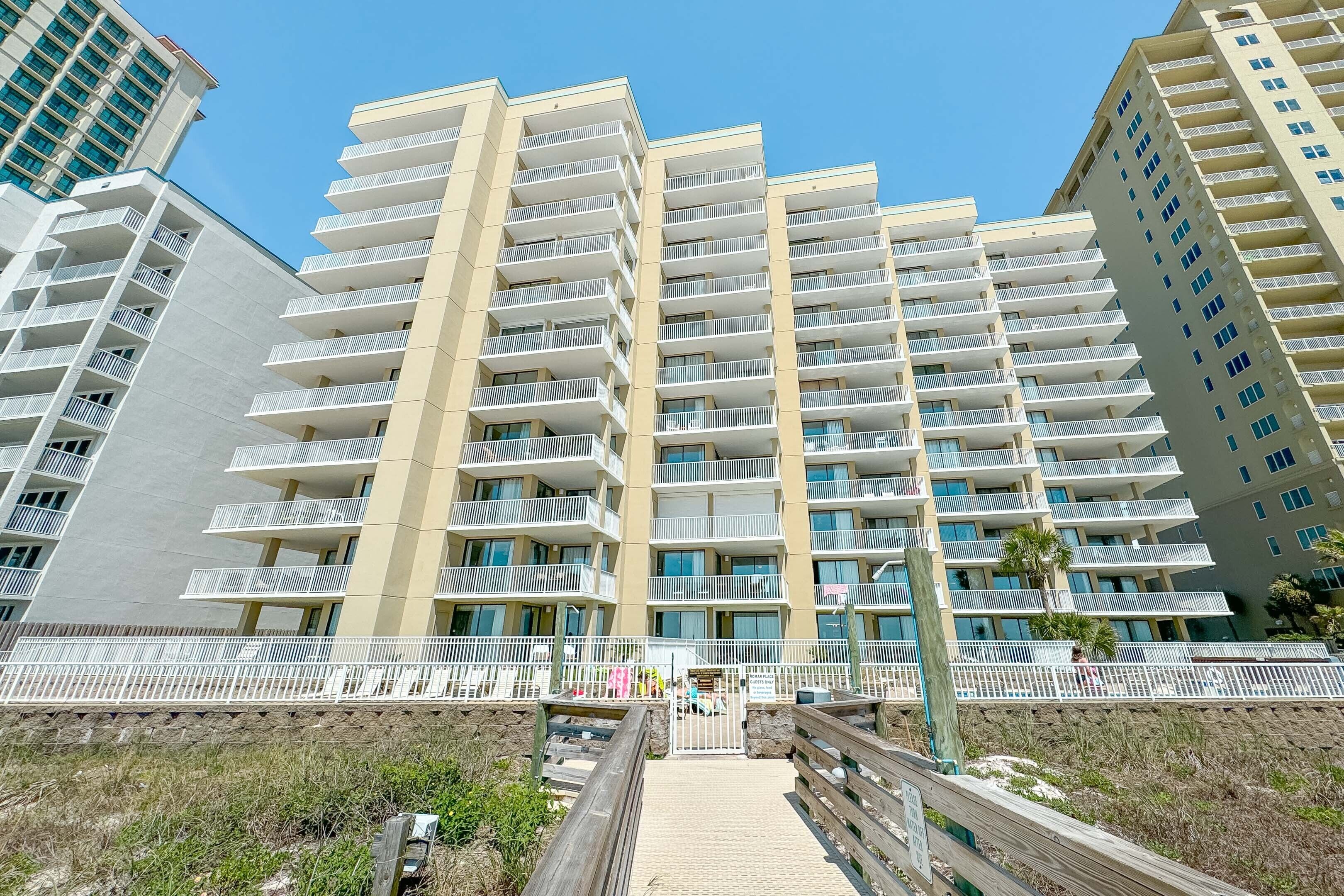 Un hôtel à plusieurs étages doté de balcons, situé en bord de mer, et accessible par une promenade en bois menant à l’entrée.