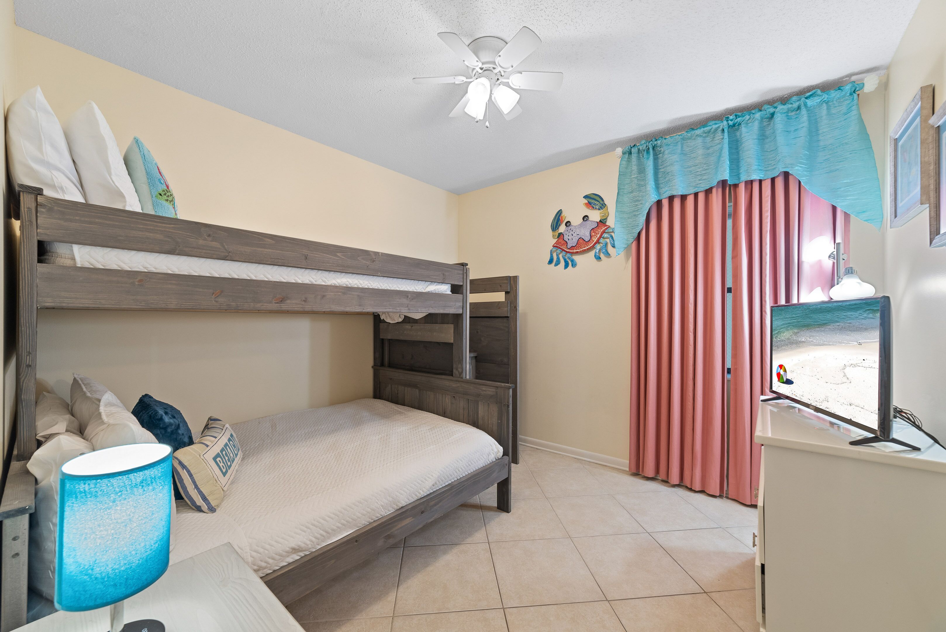 A bedroom with a bunk bed, a ceiling fan, a TV, and a window with curtains.