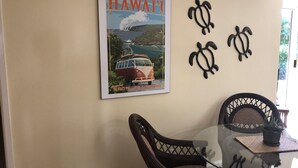 Een eethoek met een glazen tafel, twee stoelen, een poster van een oldtimer en twee schilderingen van schildpadden aan de muur.