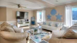Un soggiorno con un divano color beige, un divano bianco, un tavolino da caffè e una ventola a soffitto. C'è una TV a parete e stelle marine decorative.