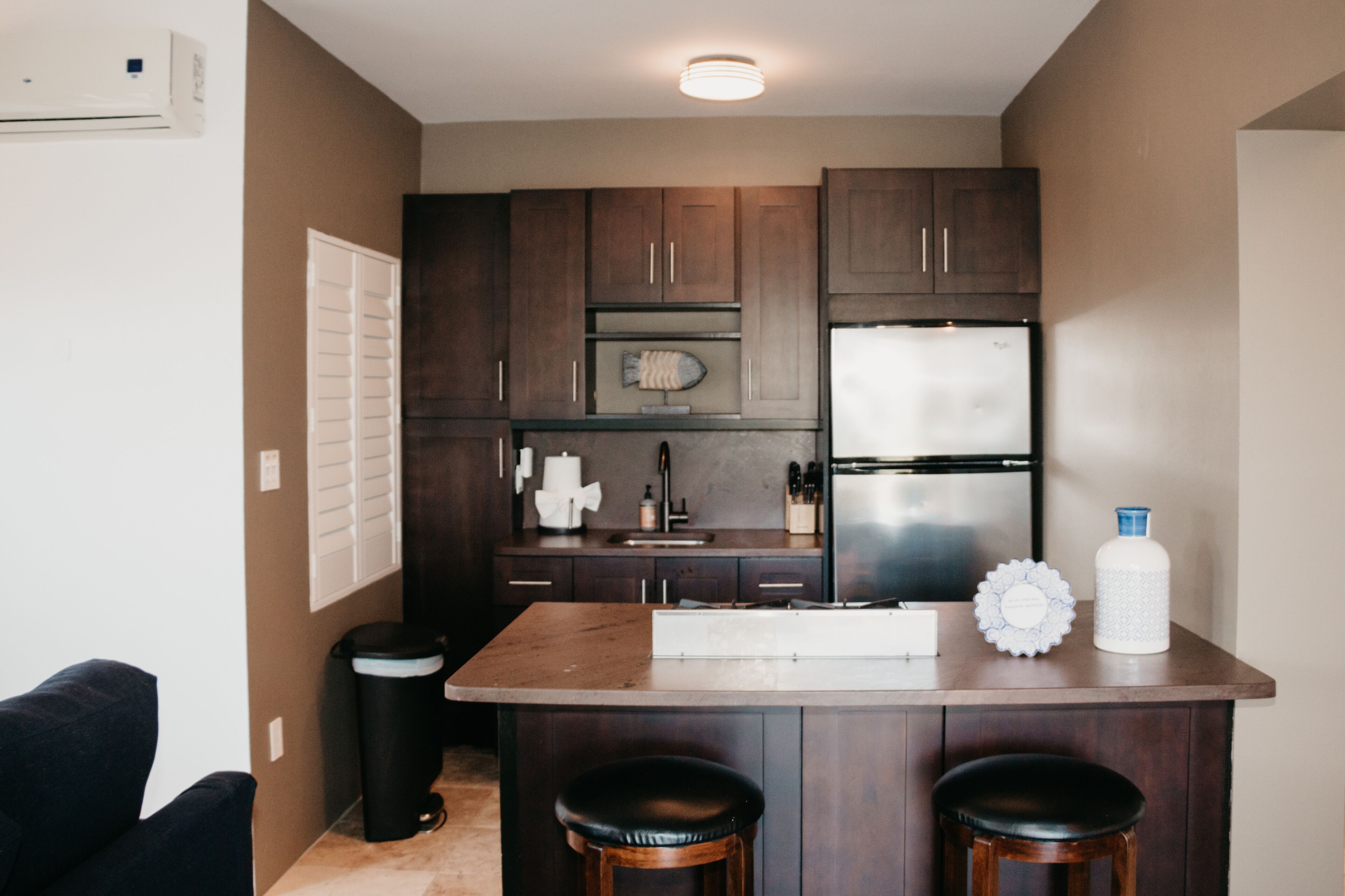 Cocina compacta con armarios de madera oscura, un refrigerador de acero inoxidable, horno empotrado y una isla central con taburetes.