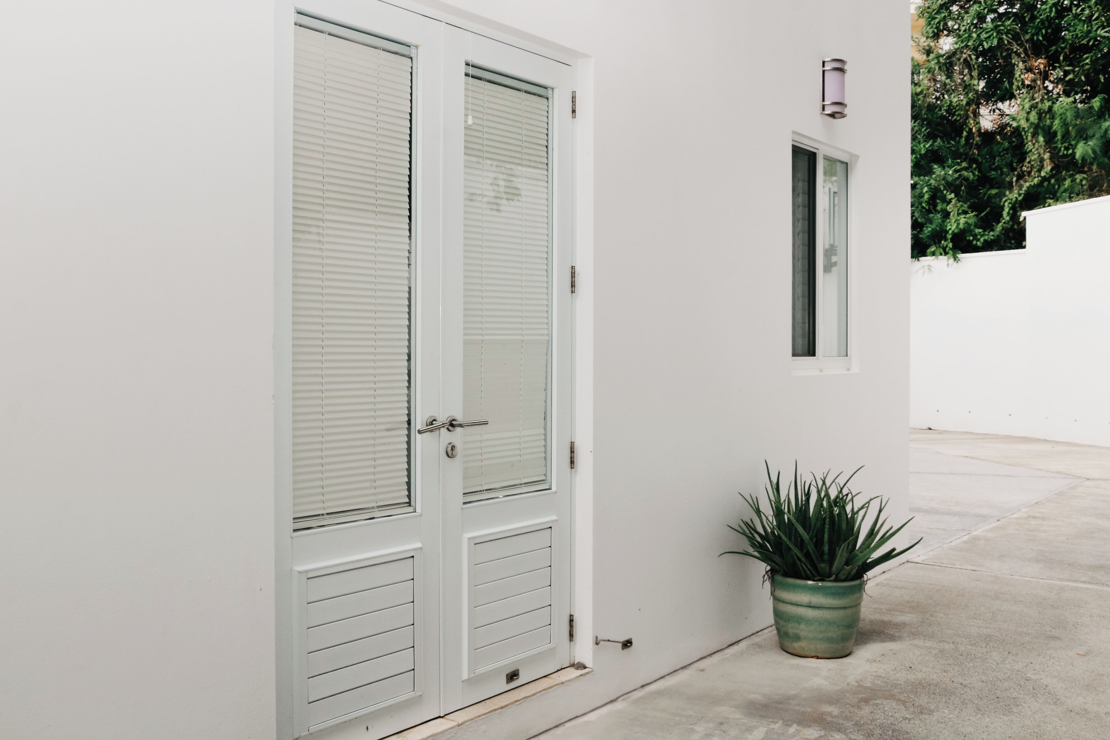 Pared exterior blanca con puerta corredera de vidrio, una ventana pequeña y una planta en maceta.