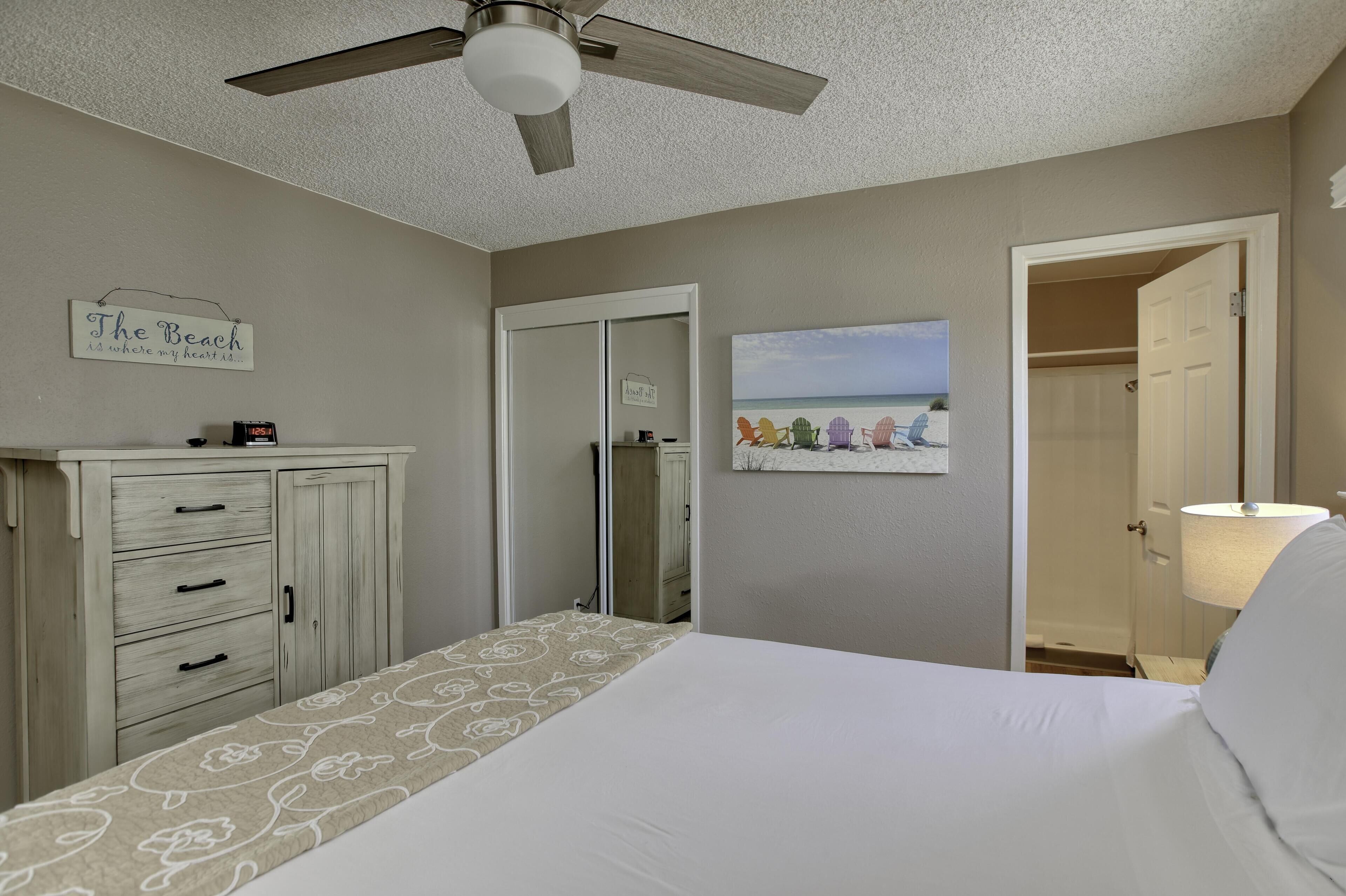 Une chambre à coucher comprenant un lit, une commode, un ventilateur de plafond, un panneau décoratif mural, un tableau encadré et un placard ouvert.