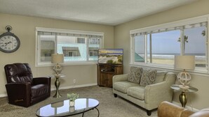 A living room with a brown leather chair, a beige sofa, a glass coffee table, and a TV on a wooden stand. There is a clock on the wall and a window with a view of the beach.