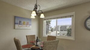 A dining area with wicker chairs, a glass table, and a painting above.