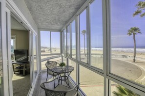 A balcony with a TV, chairs, and a table, overlooking a beach with palm trees and the ocean. - Ocean views from every room. Nothing between you and the waves. (Pismo Beach)