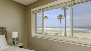 Une chambre d’hôtel avec un lit, une table de chevet avec une lampe et une fenêtre donnant sur la plage et les palmiers.