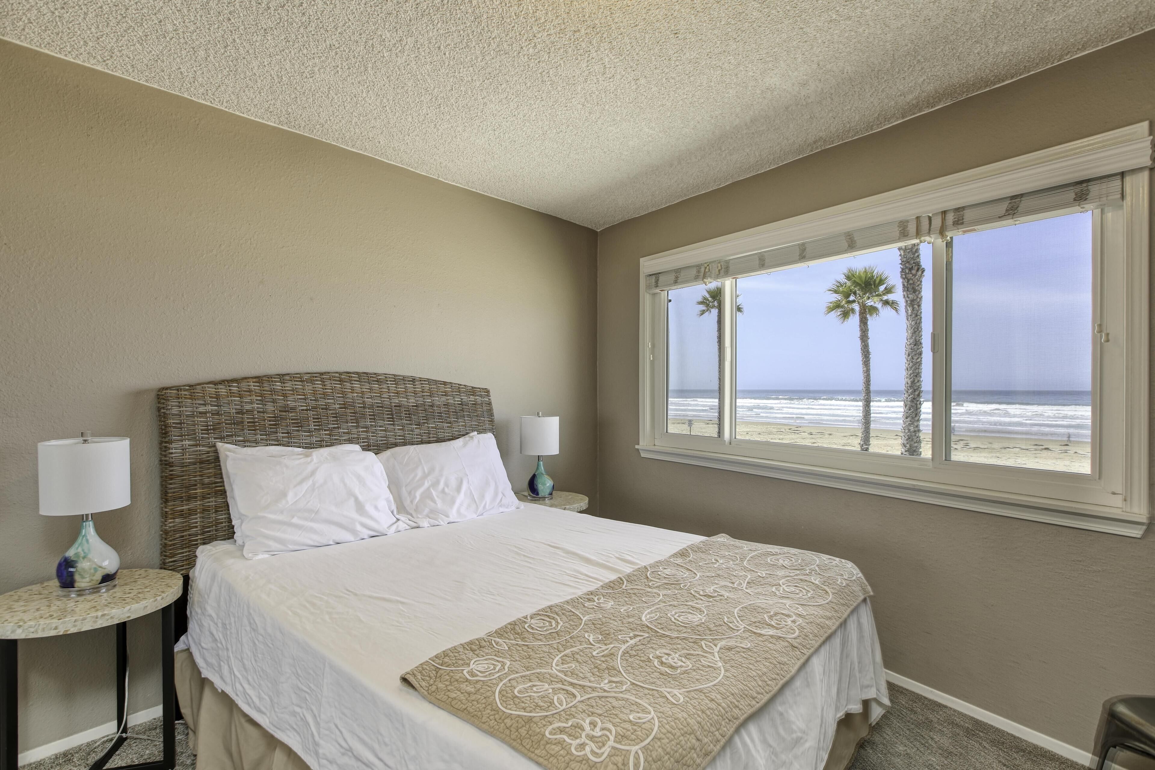 Quarto com cama, duas mesas de cabeceira com luminárias, janela com vista para a praia e palmeiras, e teto texturizado.