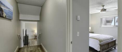 A hallway with wooden flooring, a ceiling fan, and a bed in the adjacent room.