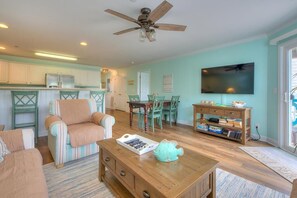 Ein Wohnzimmer mit Couchtisch aus Holz, gestreiften Sesseln, einem Essbereich mit Tisch und Stühlen, einem an der Wand montierten Fernseher und einer Küchenzeile mit Schränken und einem Kühlschrank.