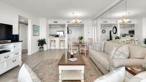 A spacious living room with a sofa, coffee table, and TV stand. There is a dining area with chairs and a chandelier. The kitchen is visible in the background. - St. Croix  #805, Awesome View From Every Room! (Destin)