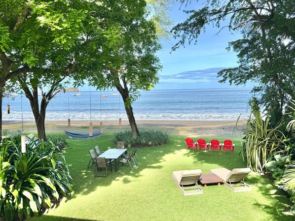 A grassy outdoor area with a table and chairs, a hammock, and red chairs facing a beach with boats in the background. - Beachfront Oasis in Playa Hermosa, Guanacaste (Playa Hermosa)
