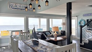 A spacious living room with a view of the ocean, featuring a wooden table, a sofa, and a staircase. - Experience oceanfront bliss at Summer Wind West in Oak Island! Ideal for 12 guests. (Oak Island)