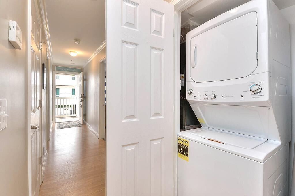 A laundry room with a front-loading washer and dryer, a white door, and a hallway leading to a balcony.