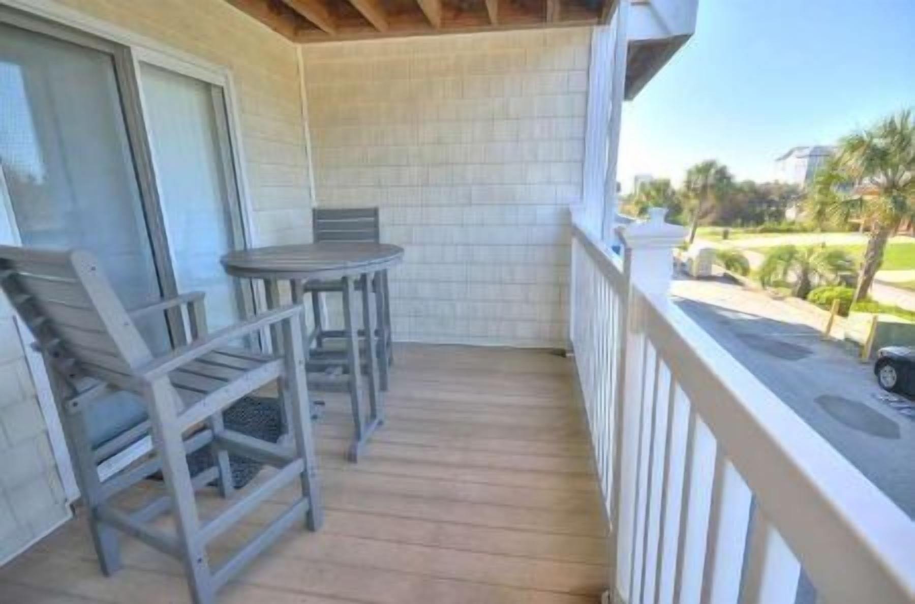 A balcony with a wooden table and two chairs, a white railing, and a view of a residential street.