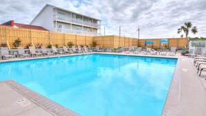 A swimming pool with lounge chairs and umbrellas, surrounded by a wooden fence and a building with balconies. - Oceanview retreat in Oak Island, 3BR condo with pool access and family amenities. (Oak Island)
