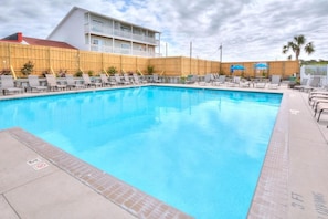A swimming pool with lounge chairs and umbrellas, surrounded by a wooden fence and a building with balconies.