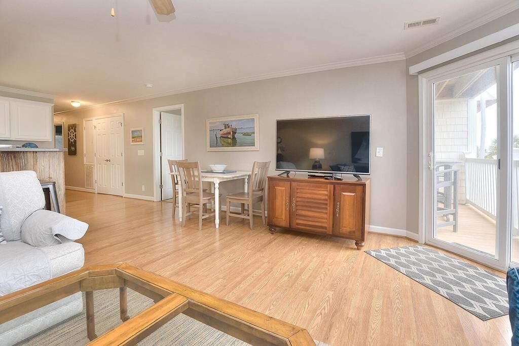 A spacious living area with a wooden floor, a dining table with chairs, a sofa, a TV, and a sliding glass door leading to a balcony.