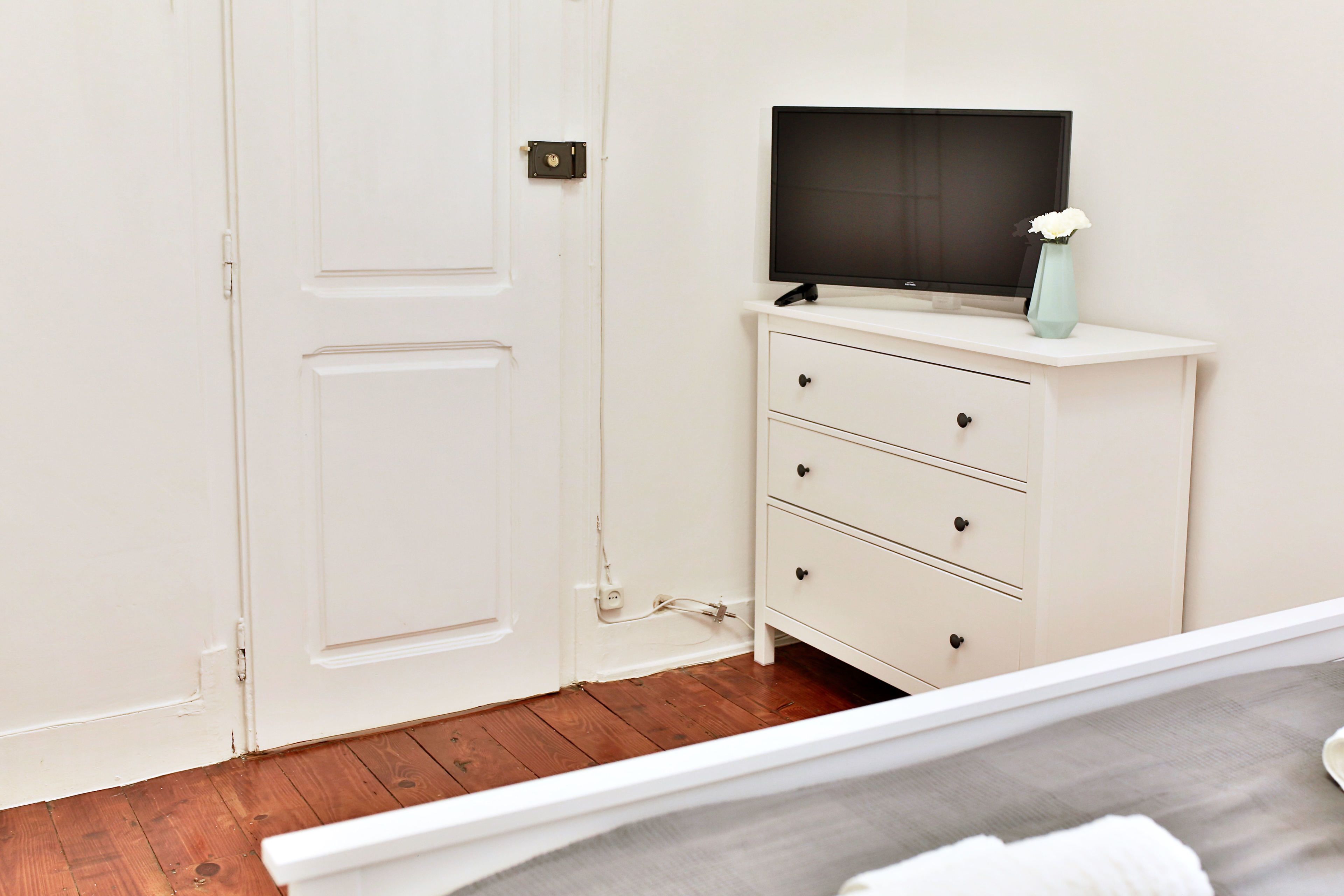 Una habitación con una puerta blanca, un aparador blanco con cajones, un televisor de pantalla plana encima y un florero con flores.
