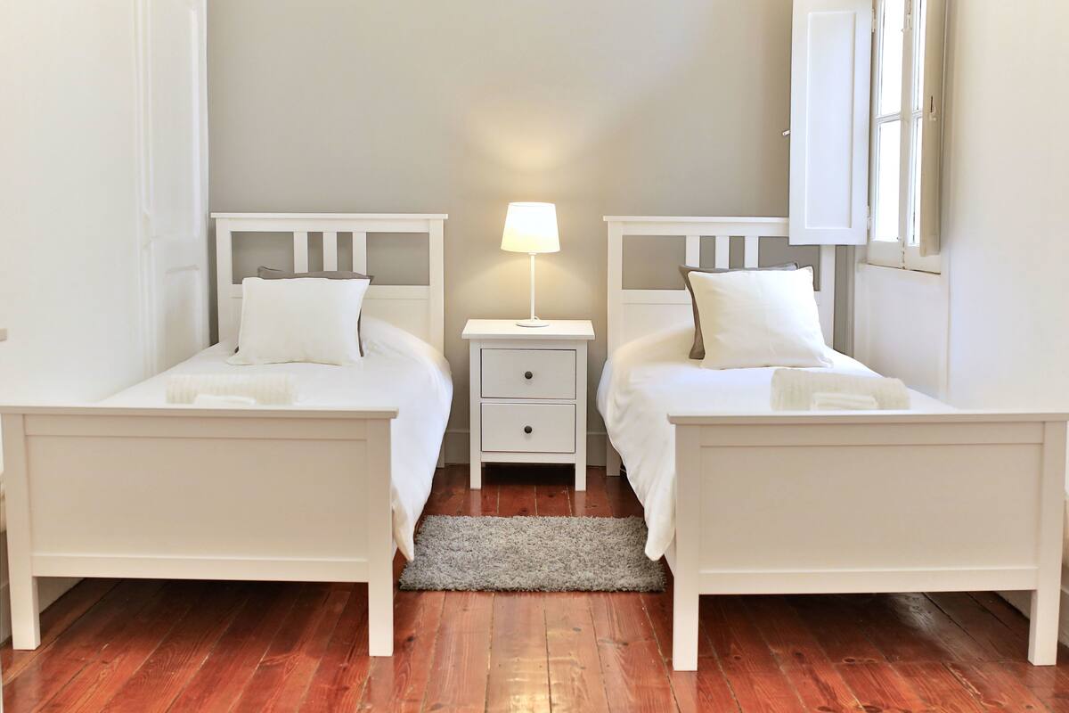 Une chambre double avec deux lits simples, une table de chevet avec une lampe, un petit tiroir et un tapis gris.