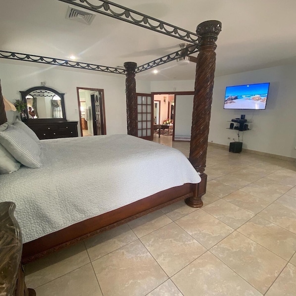 A spacious bedroom with a four-poster bed, a television mounted on the wall, and a large window.