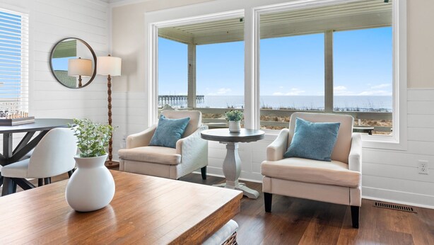 A room with a wooden table, two armchairs, a round table with a lamp, and large windows with a view of the sea. - Experience oceanfront luxury at Pier Magic, Oak Island – 6 bedrooms for up to 16 guests. (Oak Island)