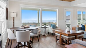 A spacious room with a wooden ceiling, large windows with a view of the beach, a round dining table, and comfortable seating. - Experience oceanfront luxury at Pier Magic, Oak Island – 6 bedrooms for up to 16 guests. (Oak Island)