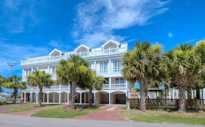A large, white, multi-story building with a balcony on the second floor, surrounded by palm trees.