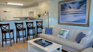A modern living room with a white sofa, a coffee table, and a kitchen area with white cabinets and a bar stool. - Escape to OW 7102-A Sweet Retreat, a 3-bedroom oceanview condo in beautiful Oak Island. (Oak Island)