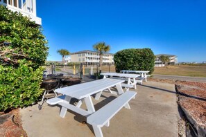 Outdoor picnic tables, a barbecue grill, and a fenced area with buildings in the background. - Escape to OW 7102-A Sweet Retreat, a 3-bedroom oceanview condo in beautiful Oak Island. (Oak Island)