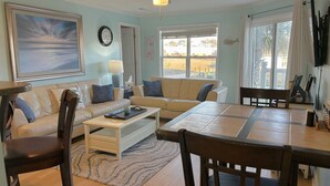 A living room with a sofa, coffee table, and dining table. There is a ceiling fan, a clock, and a window with a view of the outdoors. - Escape to OW 7102-A Sweet Retreat, a 3-bedroom oceanview condo in beautiful Oak Island. (Oak Island)