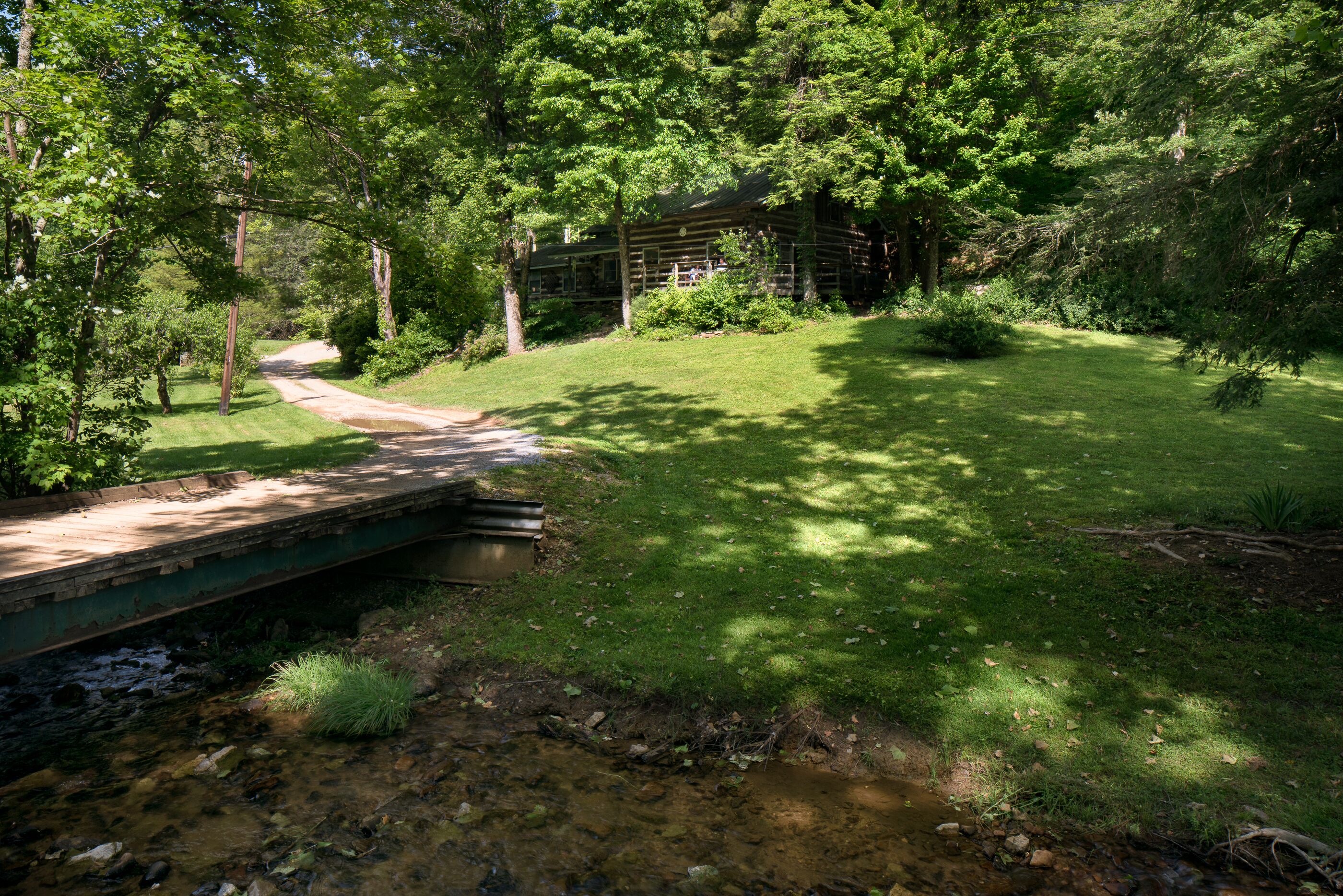 A rustic cabin surrounded by lush greenery, a wooden bridge over a stream, and a well-maintained lawn.
